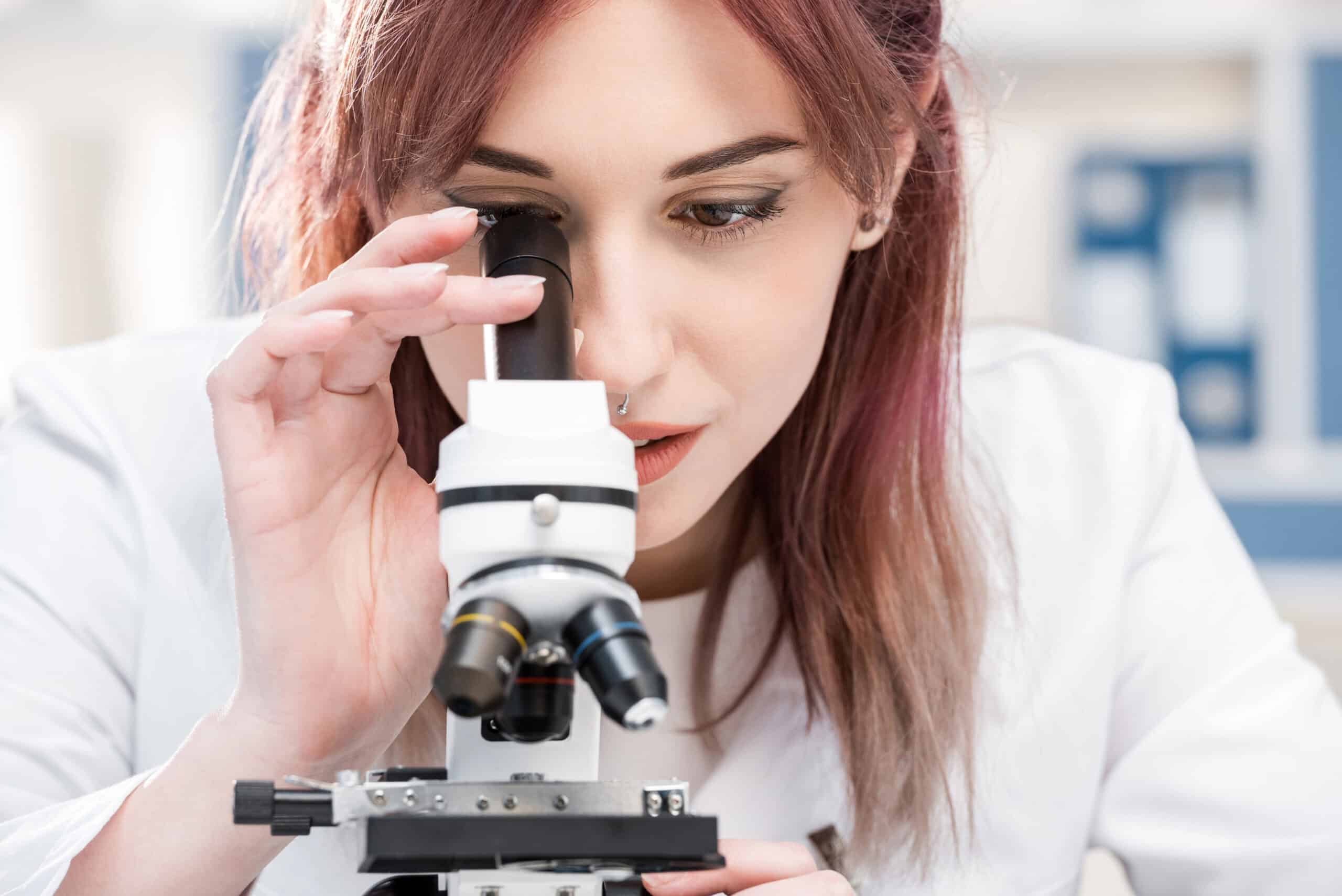 אישה צעירה בחלוק מעבדה לבן מביטה במיקרוסקופ, מתמקדת בריכוז רב תוך כדי ביצוע ניסוי מדעי או מחקר במעבדה.